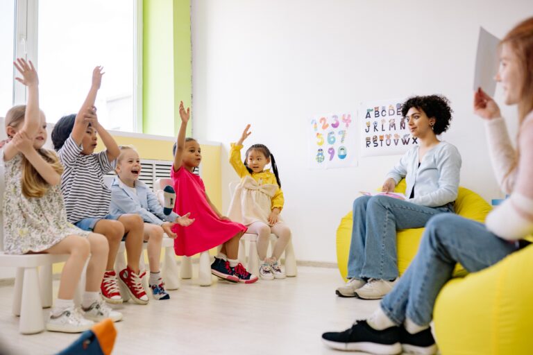 Young children raising their hands in a bright childcare classroom setting.