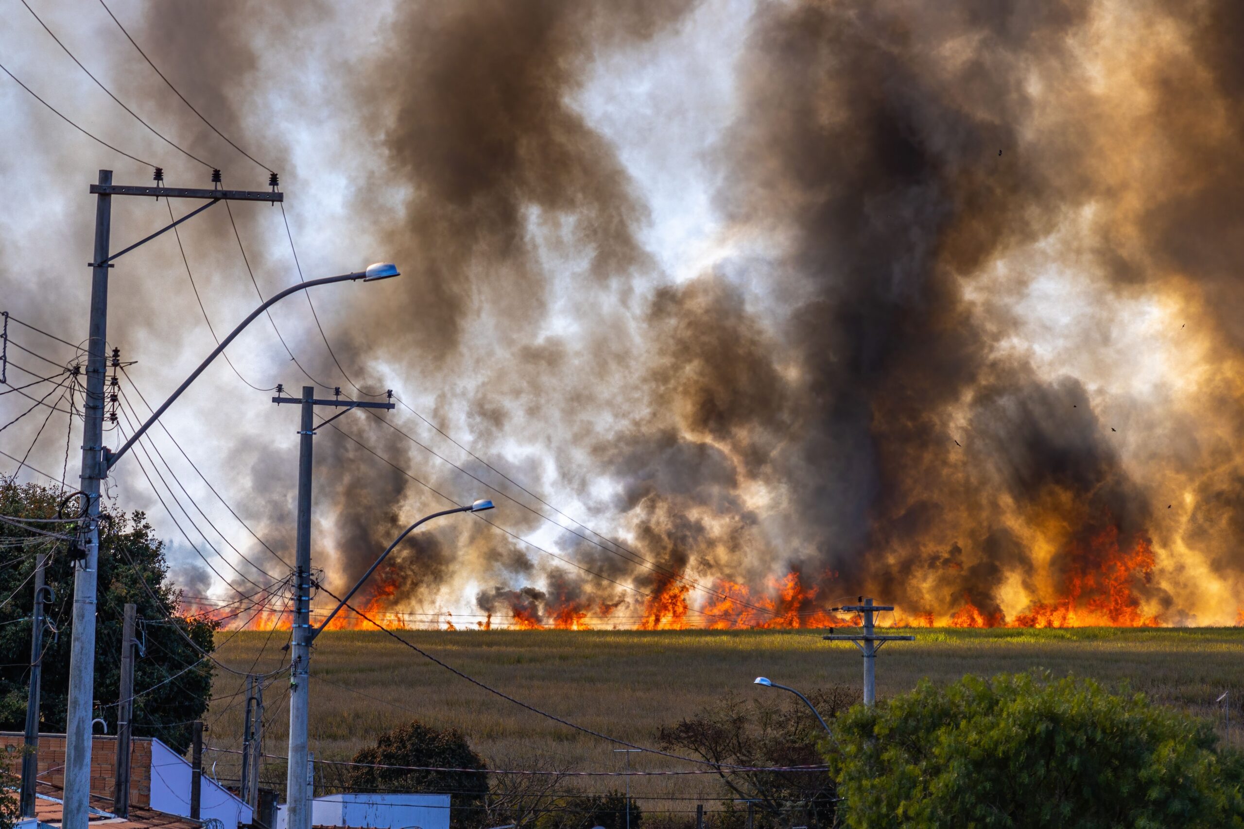 Wildfire burning across a grass field, sending thick smoke into the sky near roadside power lines